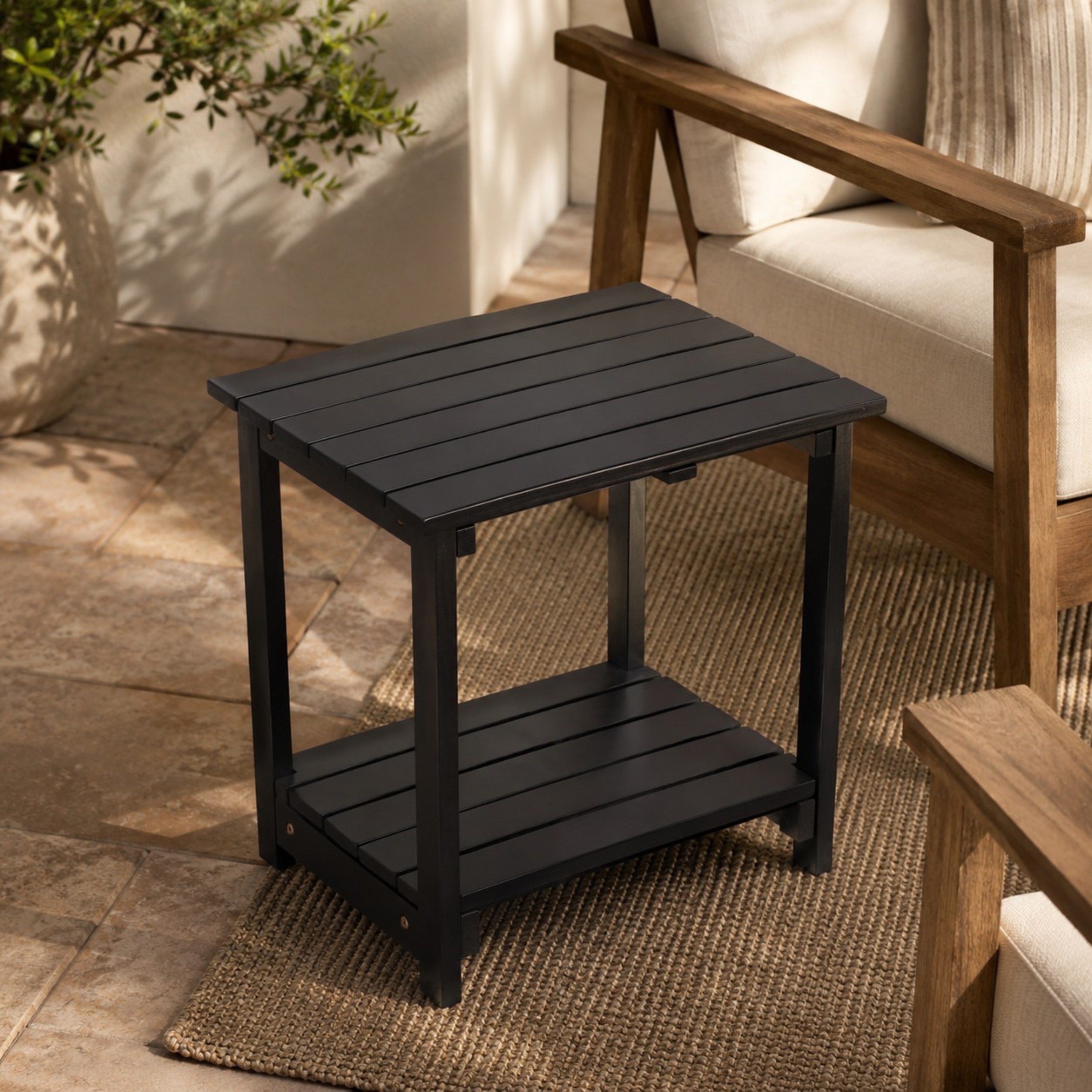 A black outdoor side table with a slatted top and lower shelf, shown in a patio setting next to a wooden armchair.