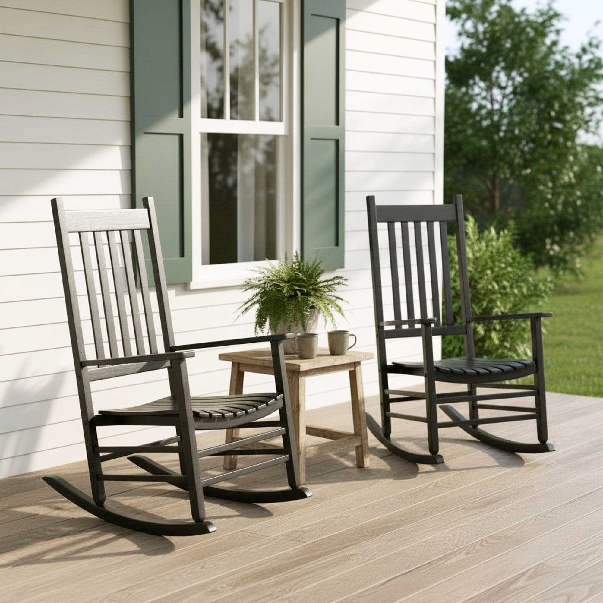 Two black rocking chairs on a wooden front porch beside a small side table with a potted plant and mug, set against a white house with green shutters.