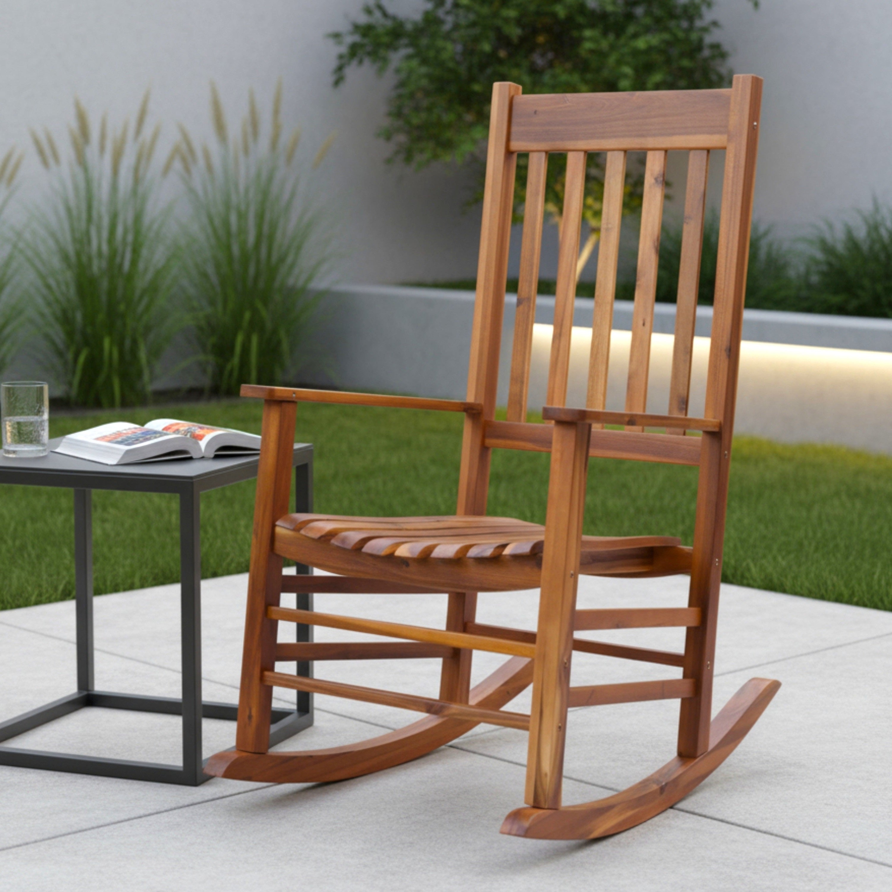 A wooden outdoor rocking chair with a slatted back and seat, shown on a patio beside a small black side table in a landscaped garden setting.
