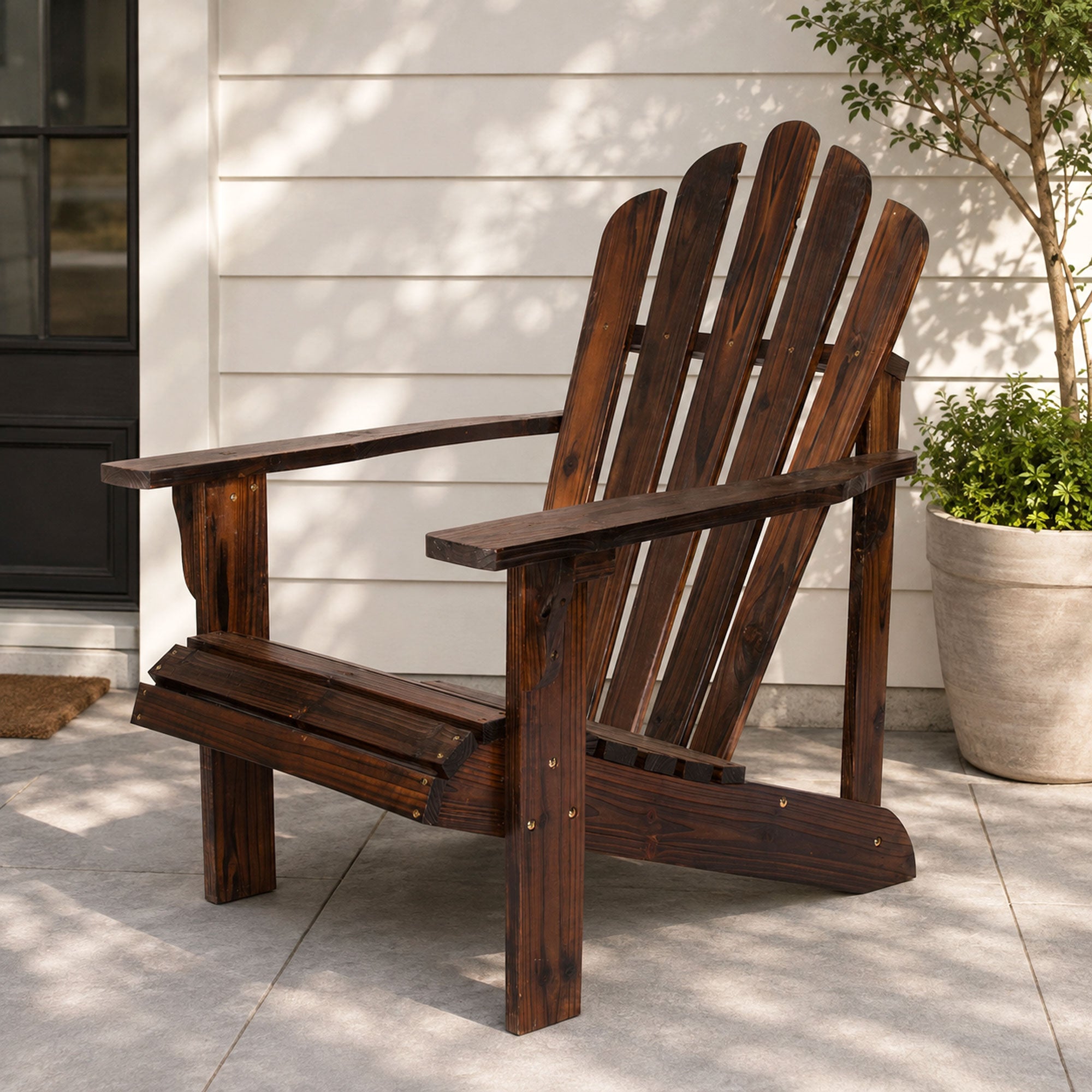 A dark brown wooden Adirondack chair placed on a light gray concrete patio in front of a white house exterior, with a potted plant to the right.