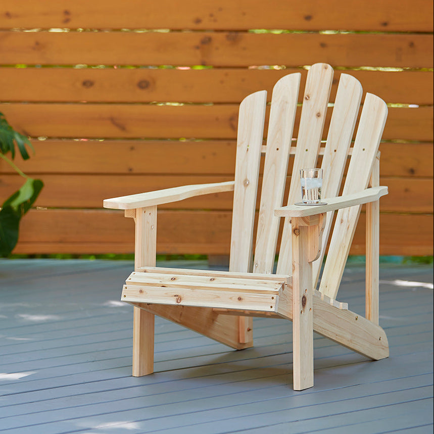 A light natural wood Adirondack chair with a small glass cup on its armrest, set on a gray wooden deck in front of a warm brown wooden fence.