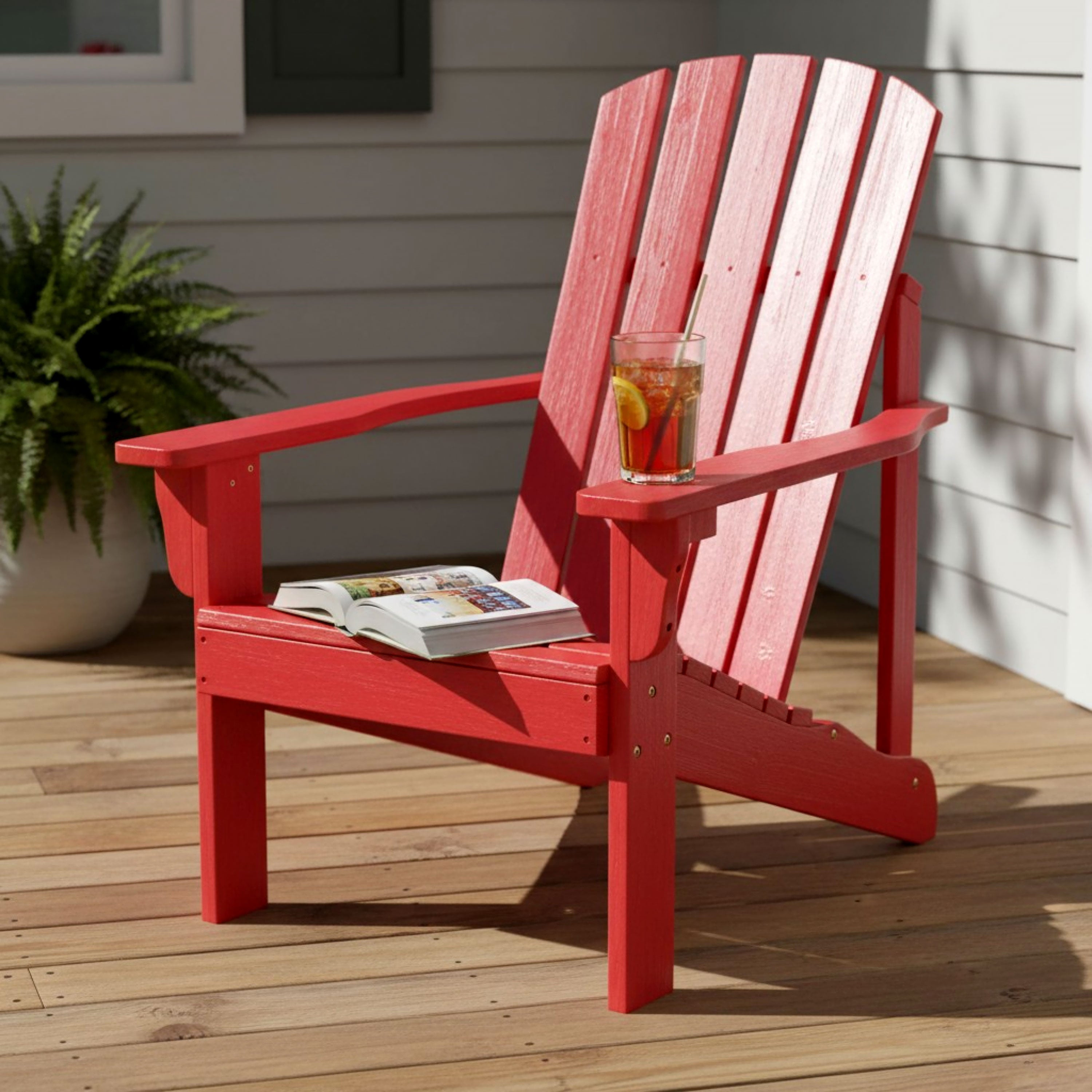 A red Adirondack-style outdoor chair on a wooden deck with a glass of iced tea on the armrest and an open magazine on the seat.