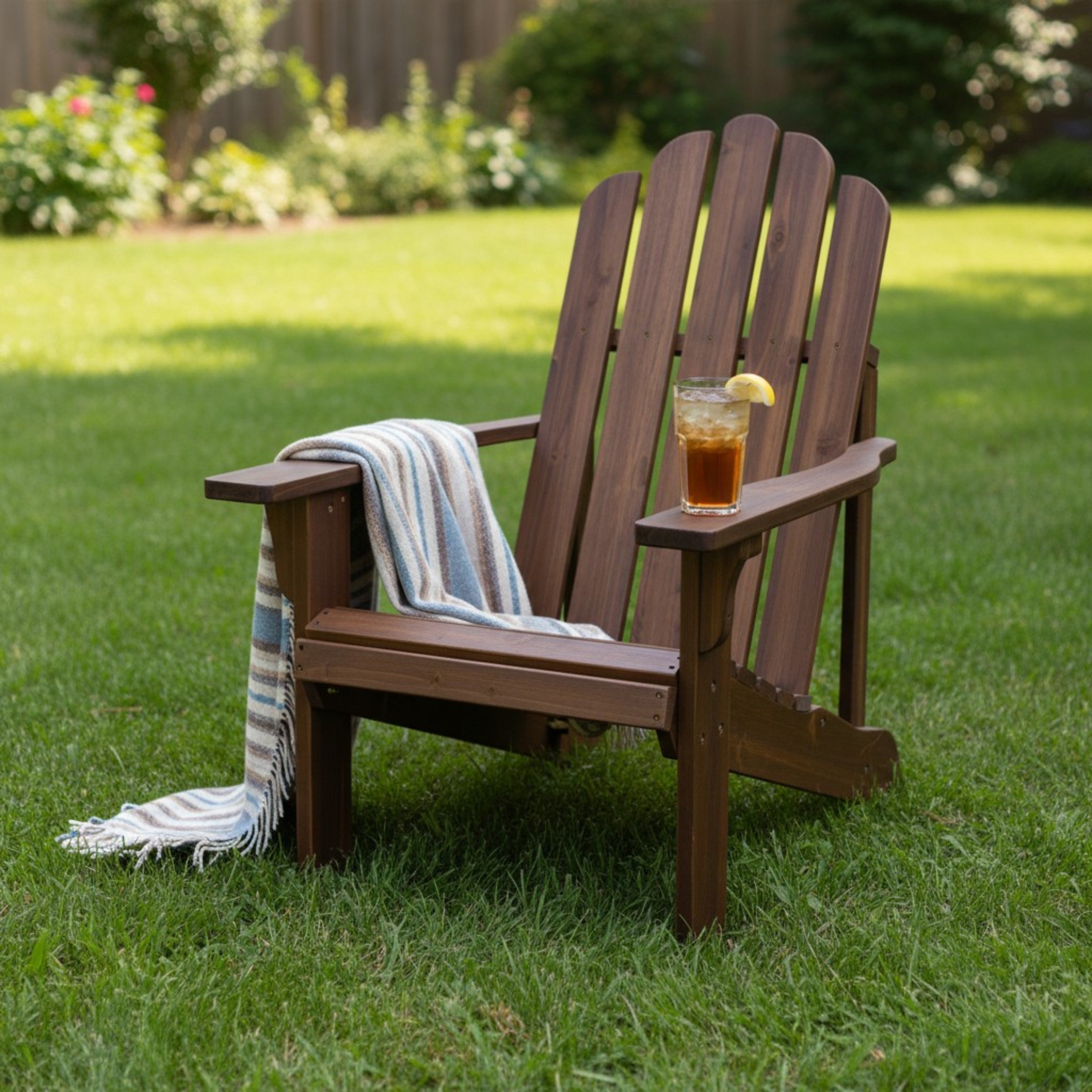 A dark brown wooden Adirondack-style outdoor chair on a green lawn, with a striped throw blanket draped over the arm and a glass of iced tea on the seat.