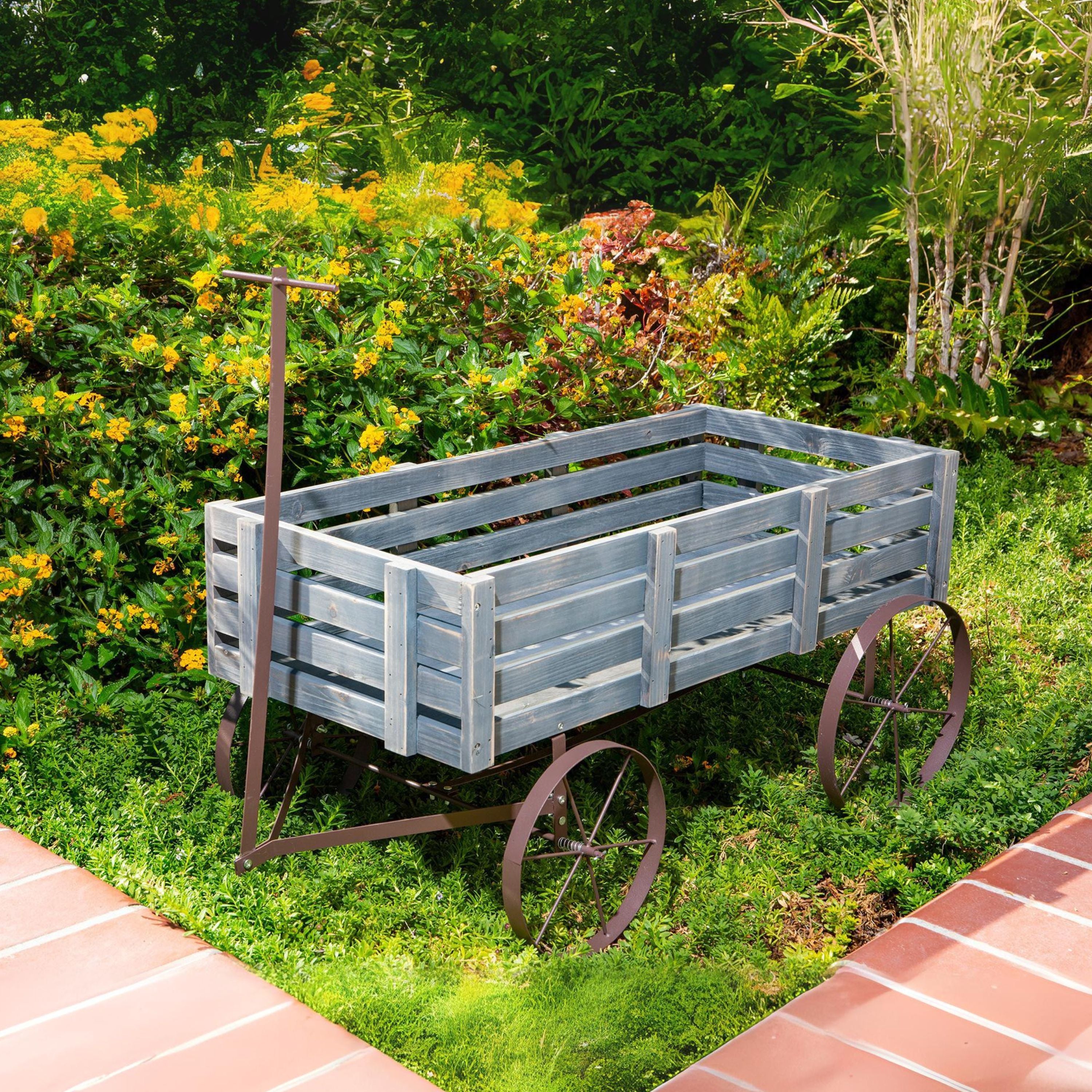A rustic gray wooden garden cart with metal wheels and a handle, placed on grass in a garden with yellow flowers and brick paths.