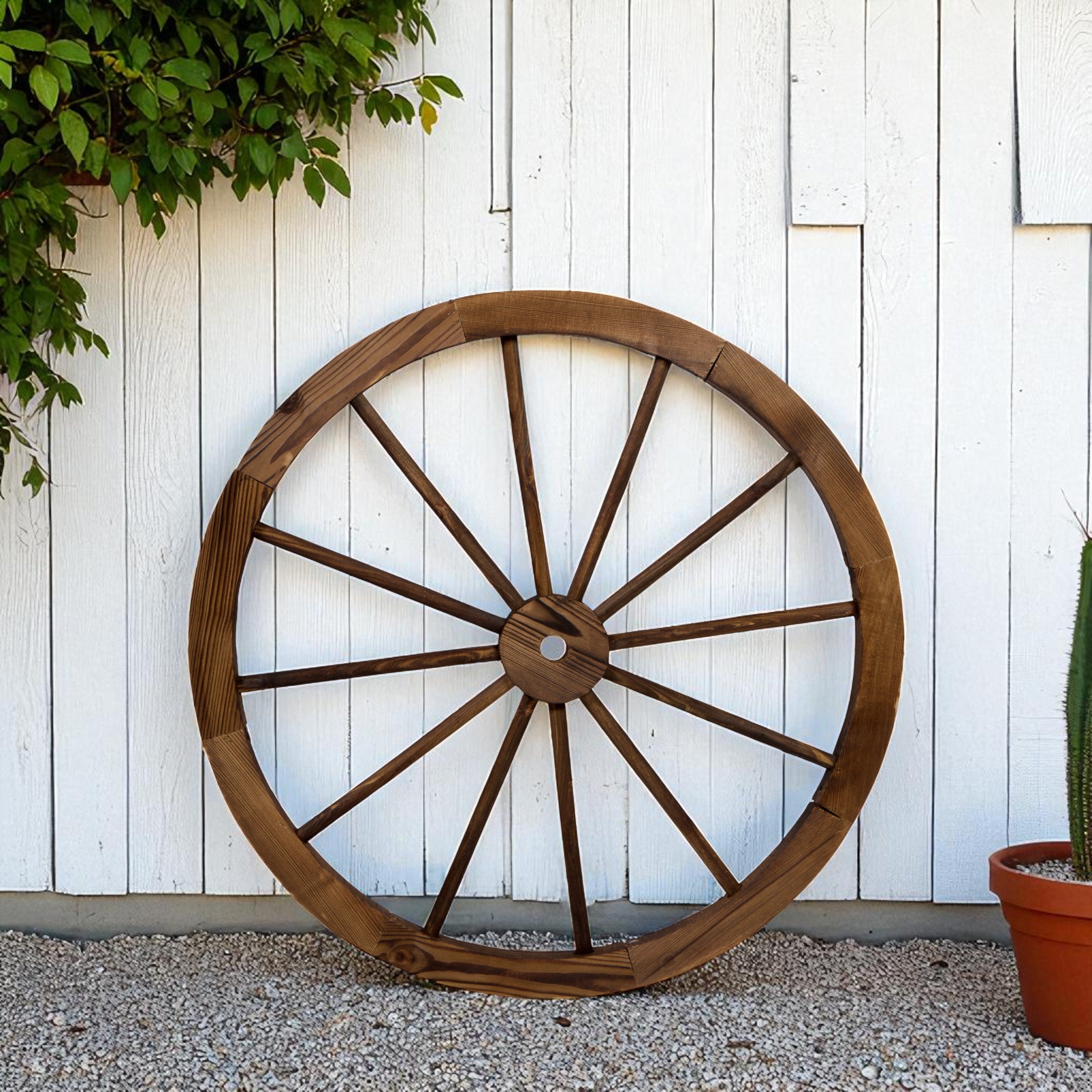 A large rustic wooden wagon wheel wall decor piece leaning against a white wood paneled wall on a gravel surface, with green foliage and a potted cactus nearby.
