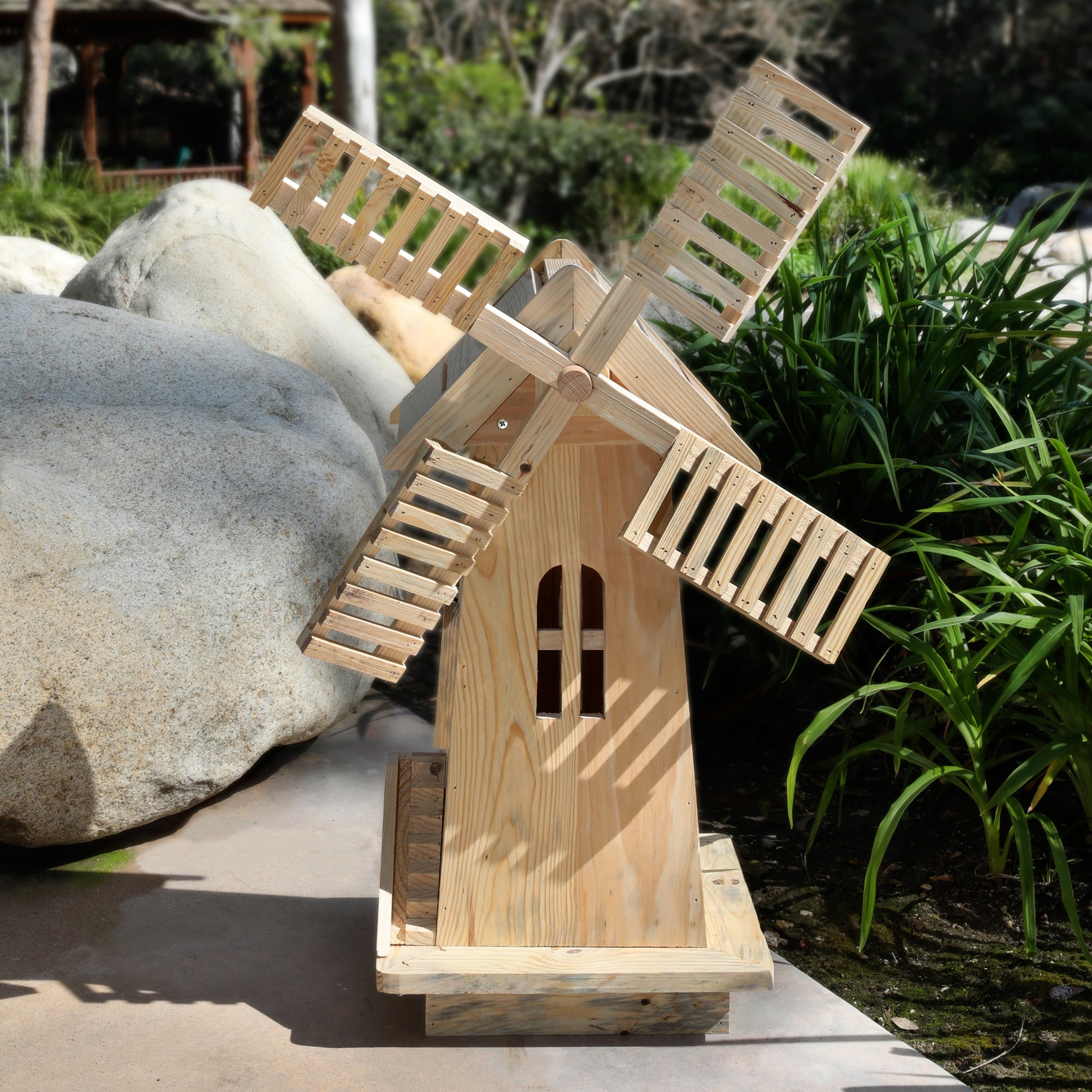 A small handcrafted wooden windmill garden ornament made from light natural wood, set outdoors on a stone surface with rocks and green plants in the background.