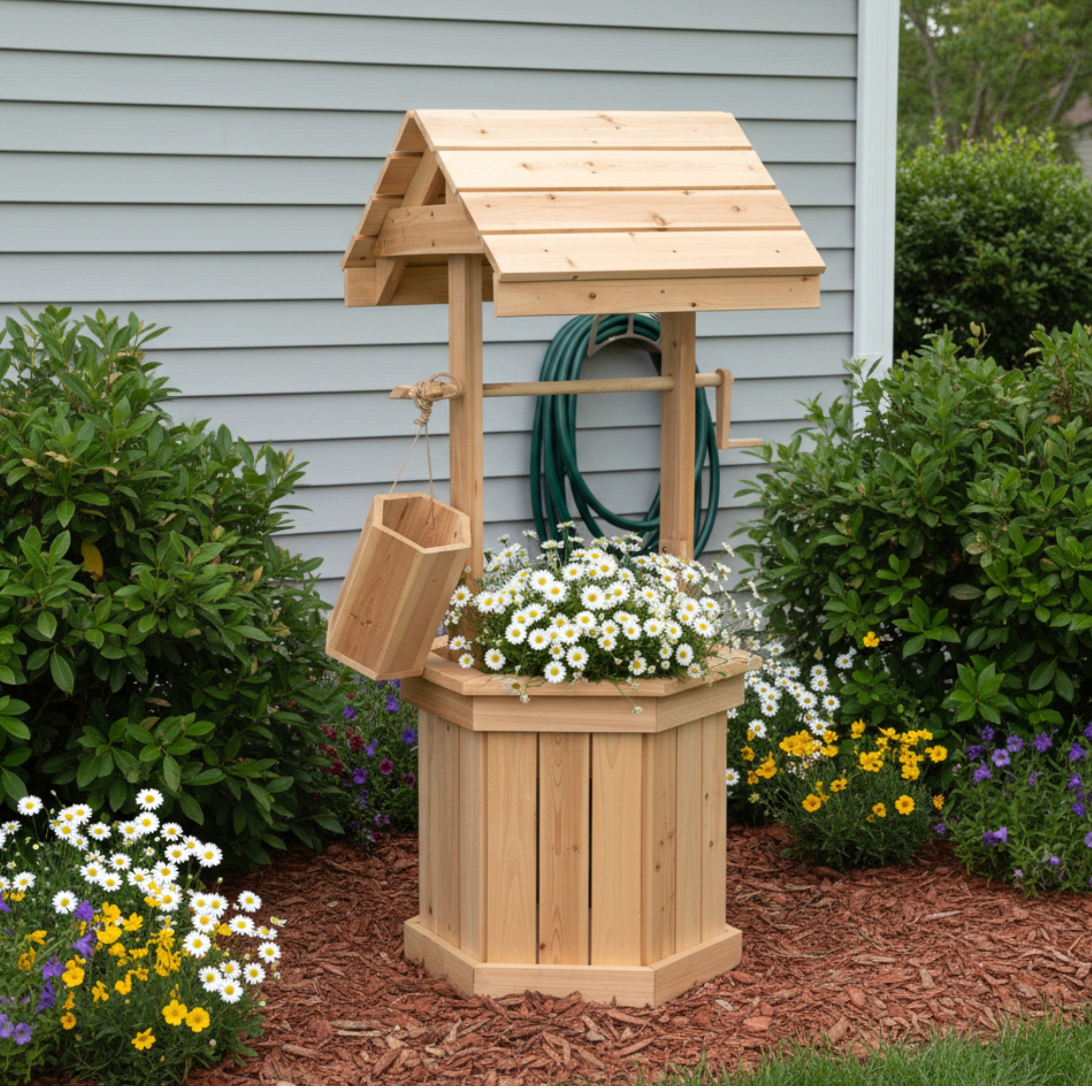 A wooden garden well planter and small well structure with a slatted bucket, shown outdoors in a landscaped garden bed in front of a gray house wall.