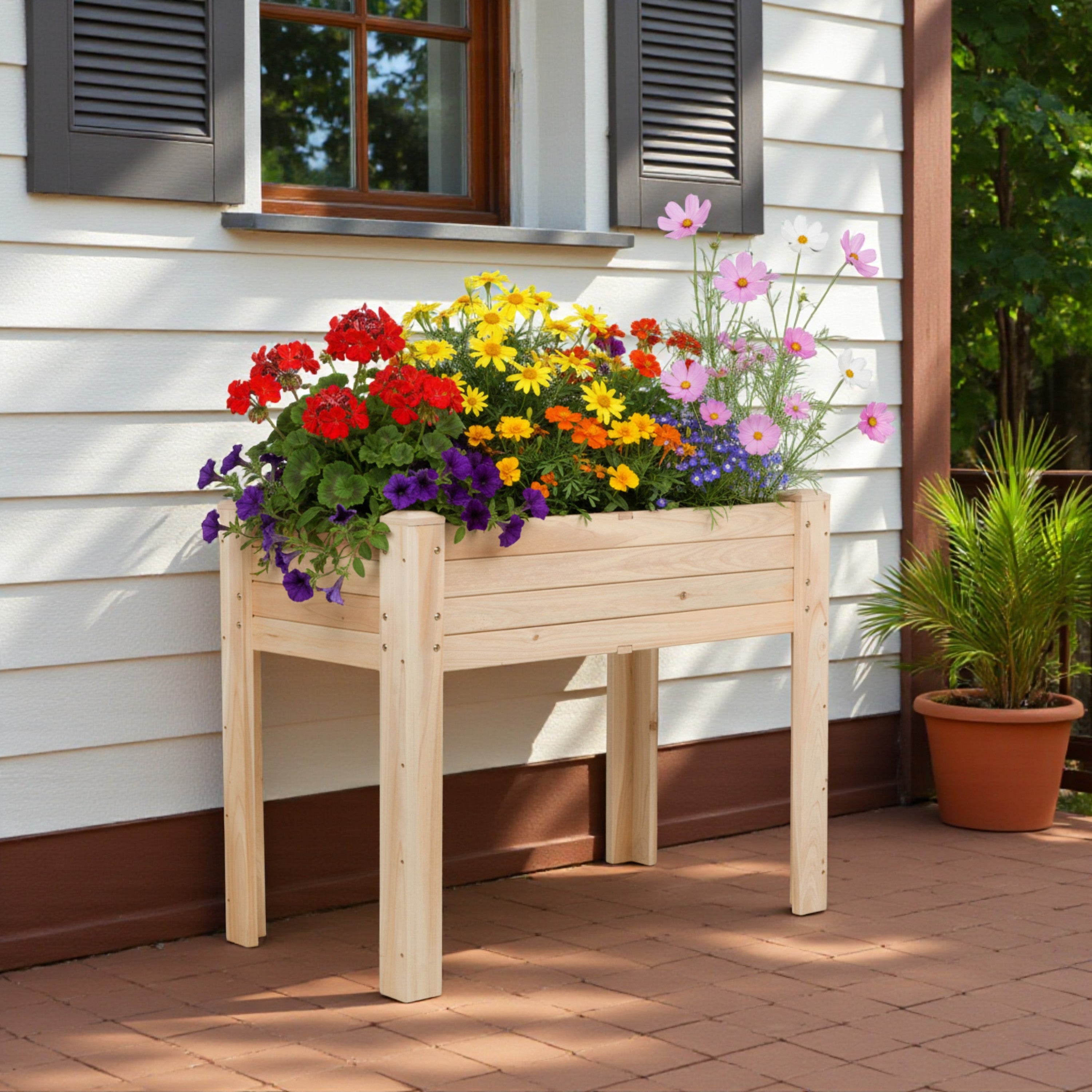 A light natural wood raised flower bed on a brick porch in front of a house with white siding and dark shutters, filled with colorful flowers including red, yellow, orange, purple, and pink blossoms.