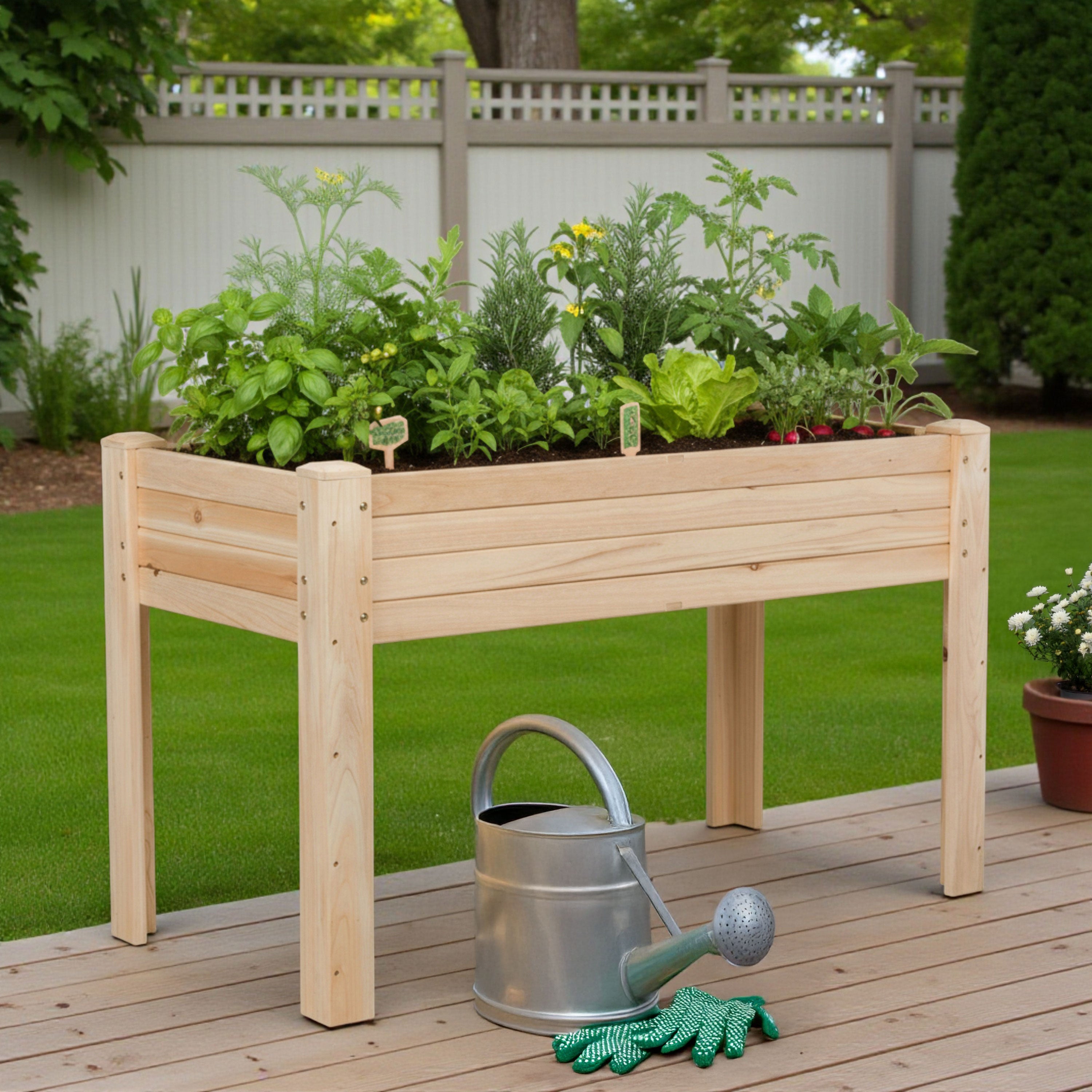 A light natural wood raised garden bed with a slatted frame, filled with various green plants, shown on a wooden deck in a backyard with a metal watering can and green gloves in front.