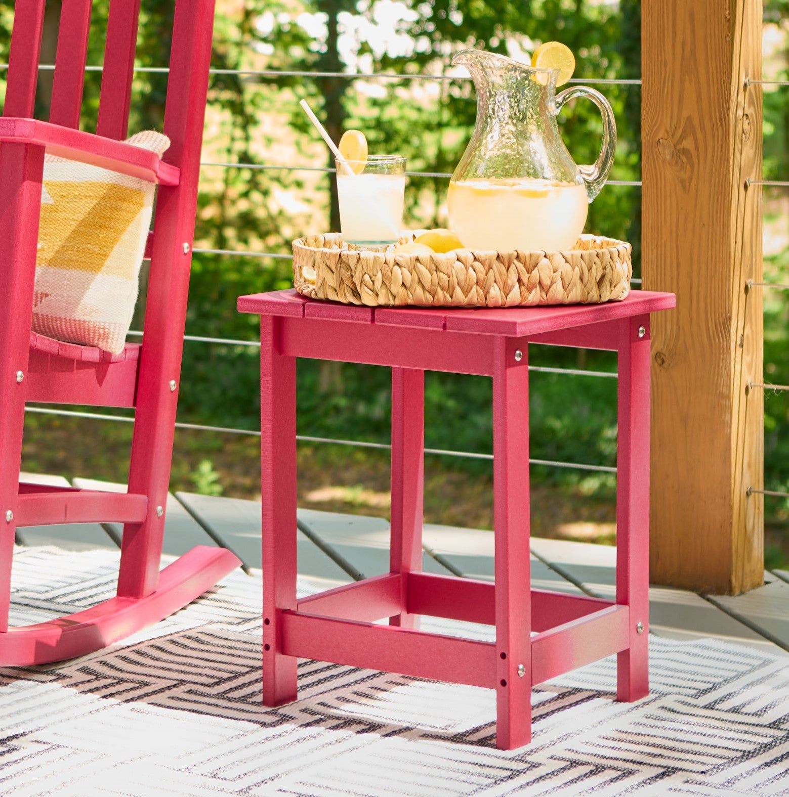 A red plastic side table on a patio, styled with a woven tray holding a glass pitcher of lemonade and a glass. A matching red rocking chair is partially visible on the left, with a green garden background.