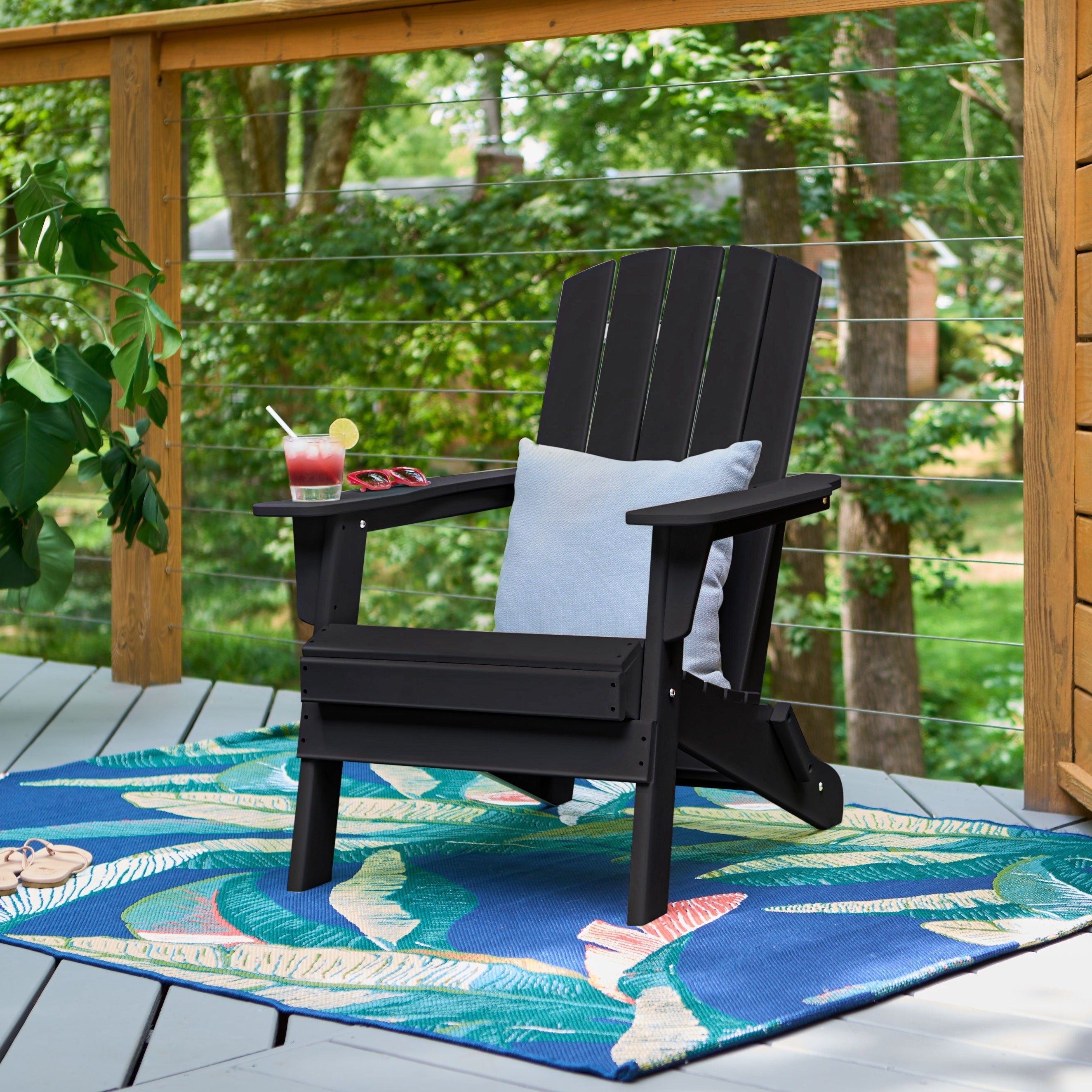 A black outdoor Adirondack chair with a white pillow sits on a blue tropical-patterned patio rug on a wooden deck, with a drink on the armrest and a wooden railing with green trees in the background.
