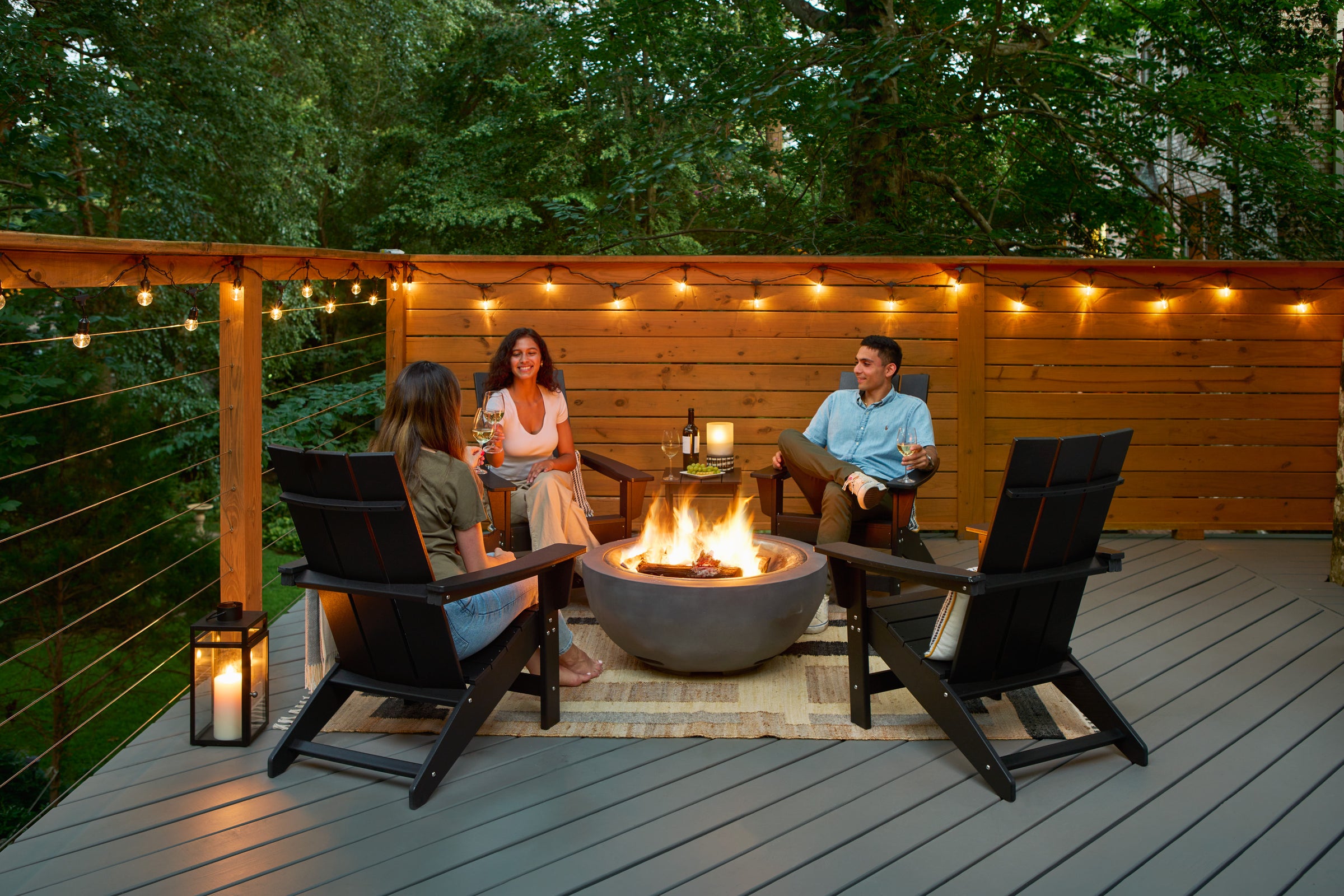 Three people sitting in black Adirondack chairs around a fire pit on a wooden deck, with string lights and a wood fence in the background.
