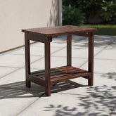 A dark brown wooden outdoor side table with a slatted top and lower shelf, shown on a light stone patio in a real outdoor setting with shadows from nearby foliage. 