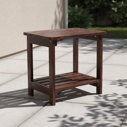 A dark brown wooden outdoor side table with a slatted top and lower shelf, shown on a light stone patio in a real outdoor setting with shadows from nearby foliage. 