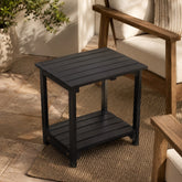 A black outdoor side table with a slatted top and lower shelf, shown in a patio setting next to a wooden armchair. 