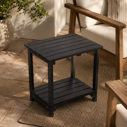 A black outdoor side table with a slatted top and lower shelf, shown in a patio setting next to a wooden armchair. 