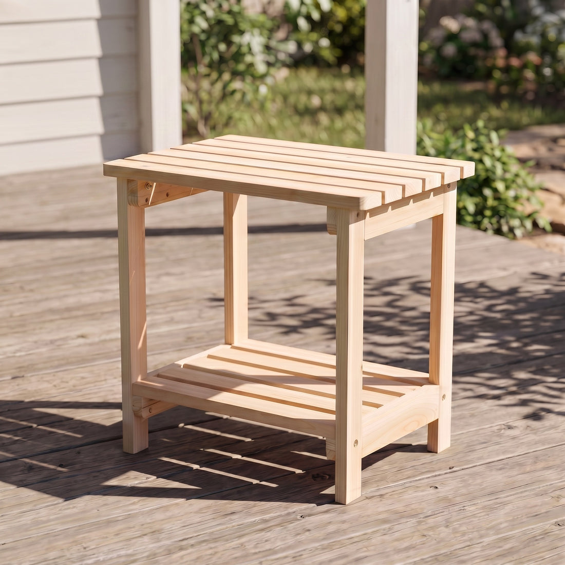 A small natural wood outdoor side table with a slatted top and lower shelf, shown on a wooden deck in a backyard setting. 