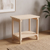 A light natural wood side table with an open lower shelf, shown in a styled room setting with a textured wall, wood floor, and a plant in the background. 