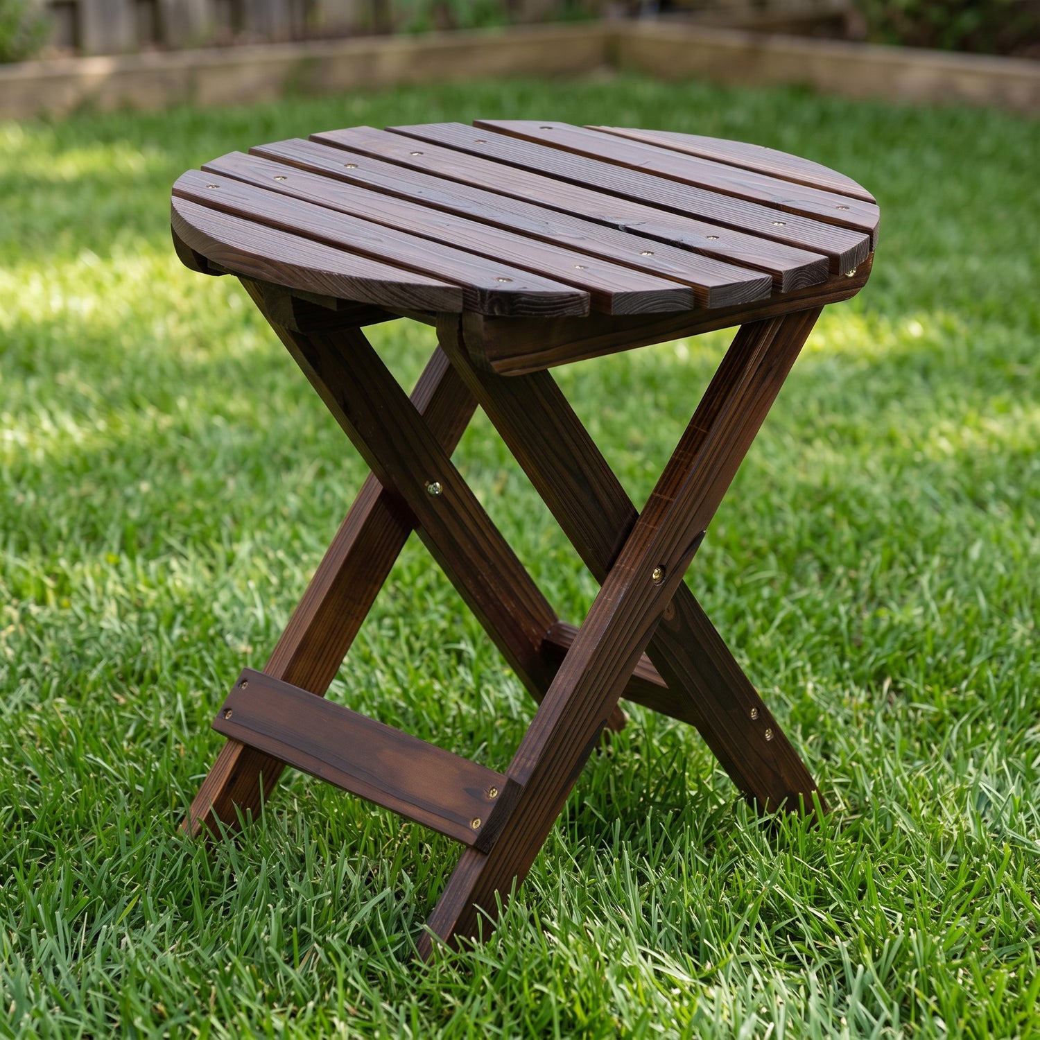 A round folding wooden side table with a dark brown slatted top and X-frame legs, shown on a green grass lawn. 