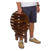 A person holding a round, foldable wooden slat table or stool against a plain white background. The person is wearing a white t-shirt, navy blue shorts, white socks, and tan lace-up shoes. 
