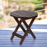 A round folding wooden side table with a dark brown slatted top and X-frame legs, shown on a blue deck with a blurred outdoor background. 