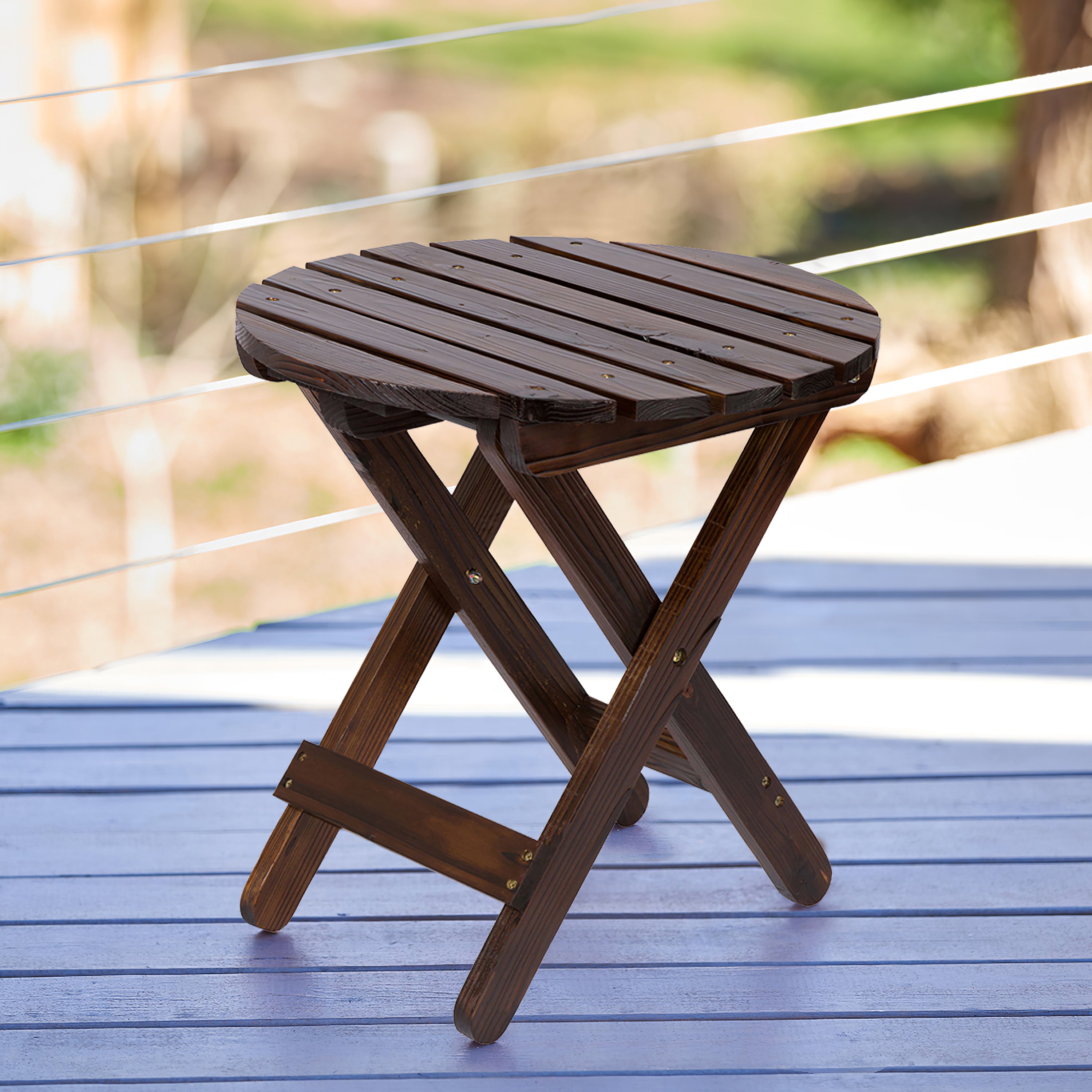 A round folding wooden side table with a dark brown slatted top and X-frame legs, shown on a blue deck with a blurred outdoor background. 