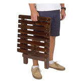 A person holding a dark brown wooden folding side table with a slatted tabletop, shown from the waist down against a plain white background. 