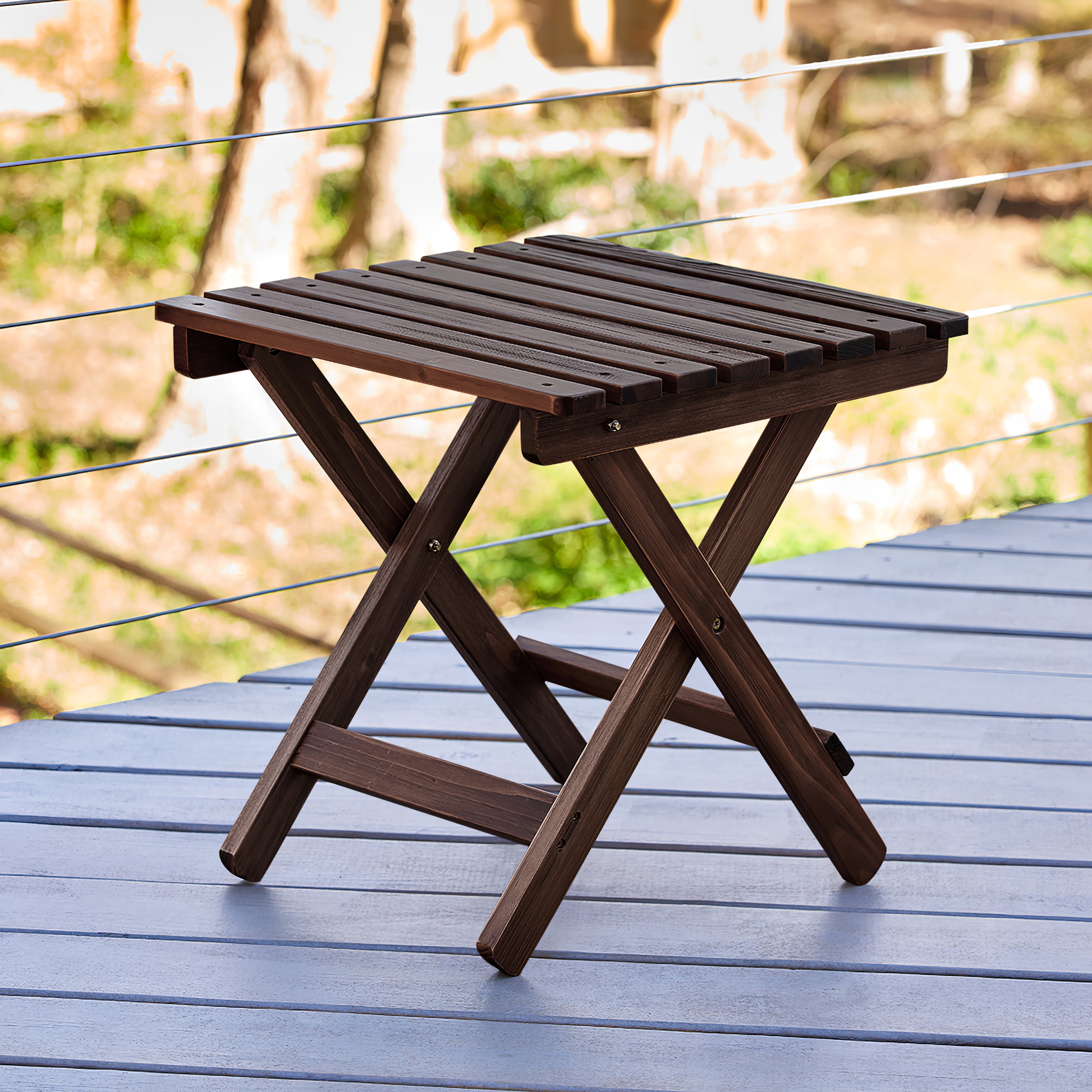 A dark brown wooden folding side table with a slatted top and X-frame legs, shown on a blue-gray deck with a blurred outdoor background. 
