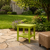 A bright lime green wooden side table with a lower shelf, placed on a stone patio in a landscaped garden setting next to a wicker chair. 
