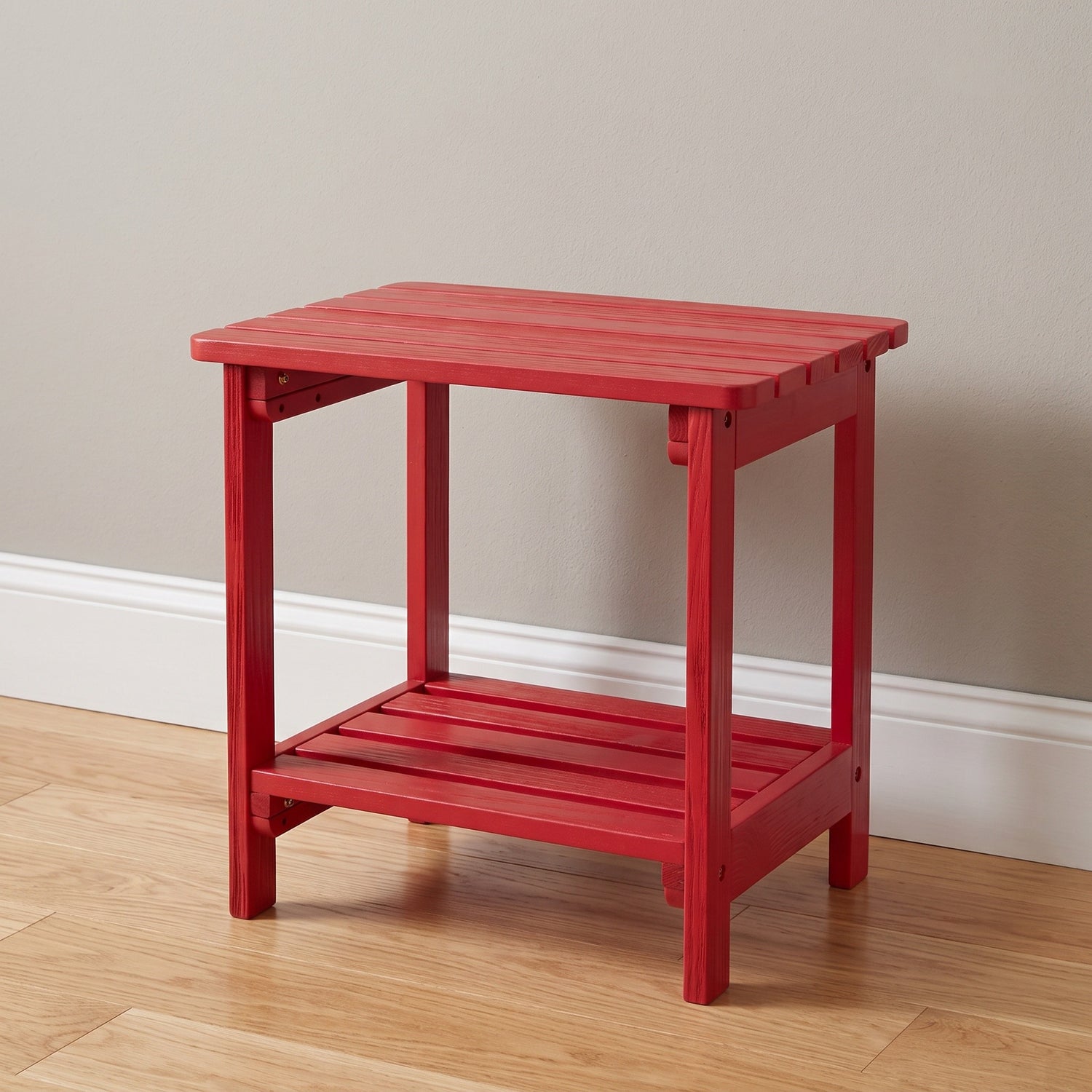 A bright red wooden end table with a slatted top and lower shelf, shown on a light wood floor against a beige wall. 