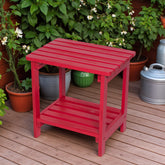 A bright red wooden side table with a slatted top and lower shelf, placed on a wooden deck with potted plants in the background. 