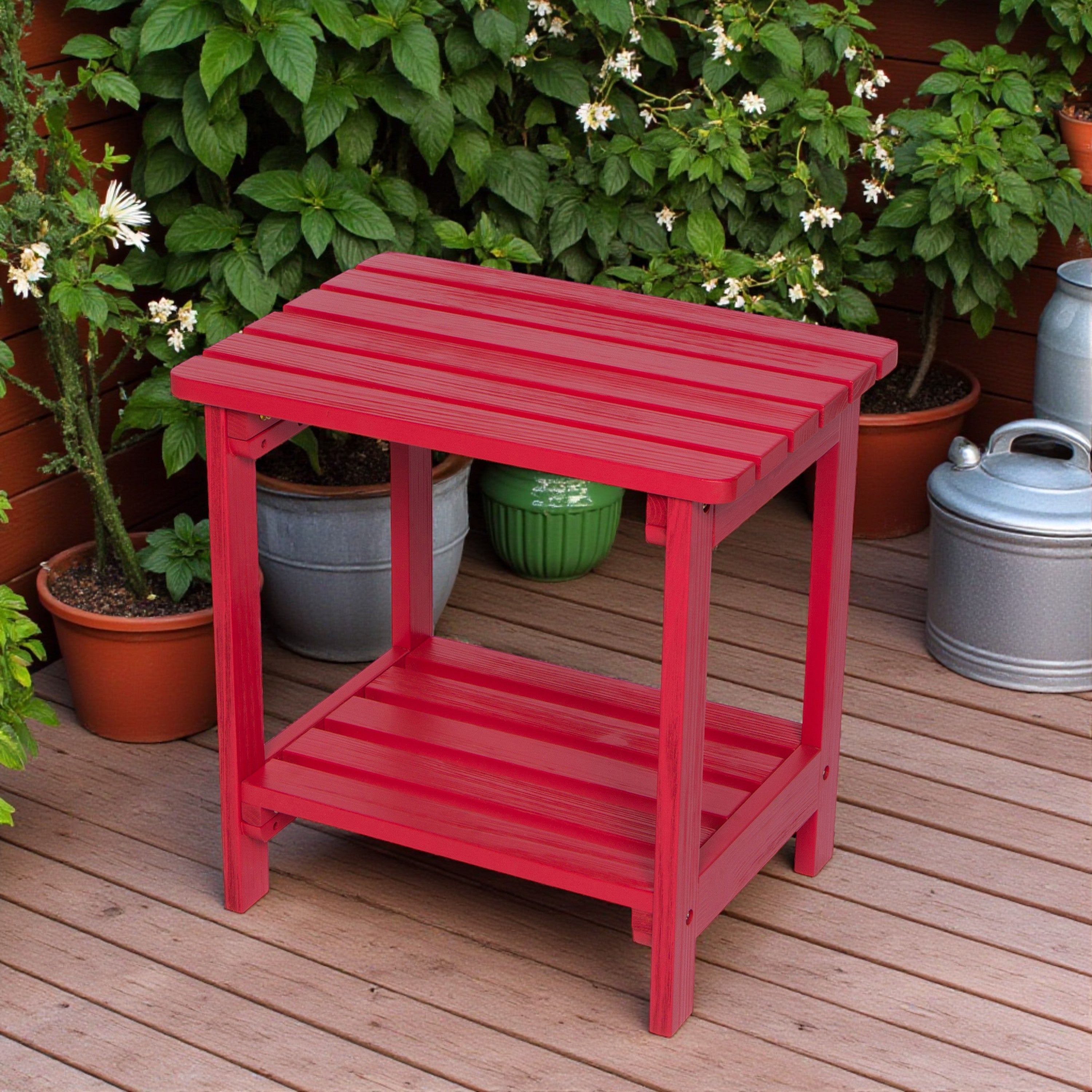 A bright red wooden side table with a slatted top and lower shelf, placed on a wooden deck with potted plants in the background. 