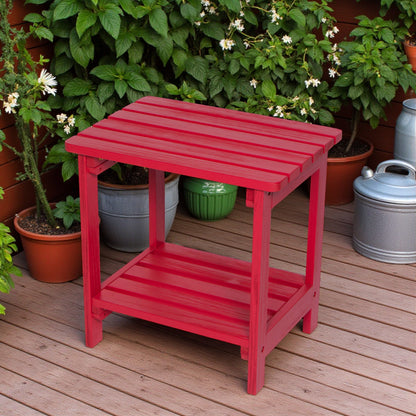 A bright red wooden side table with a slatted top and lower shelf, placed on a wooden deck with potted plants in the background. 