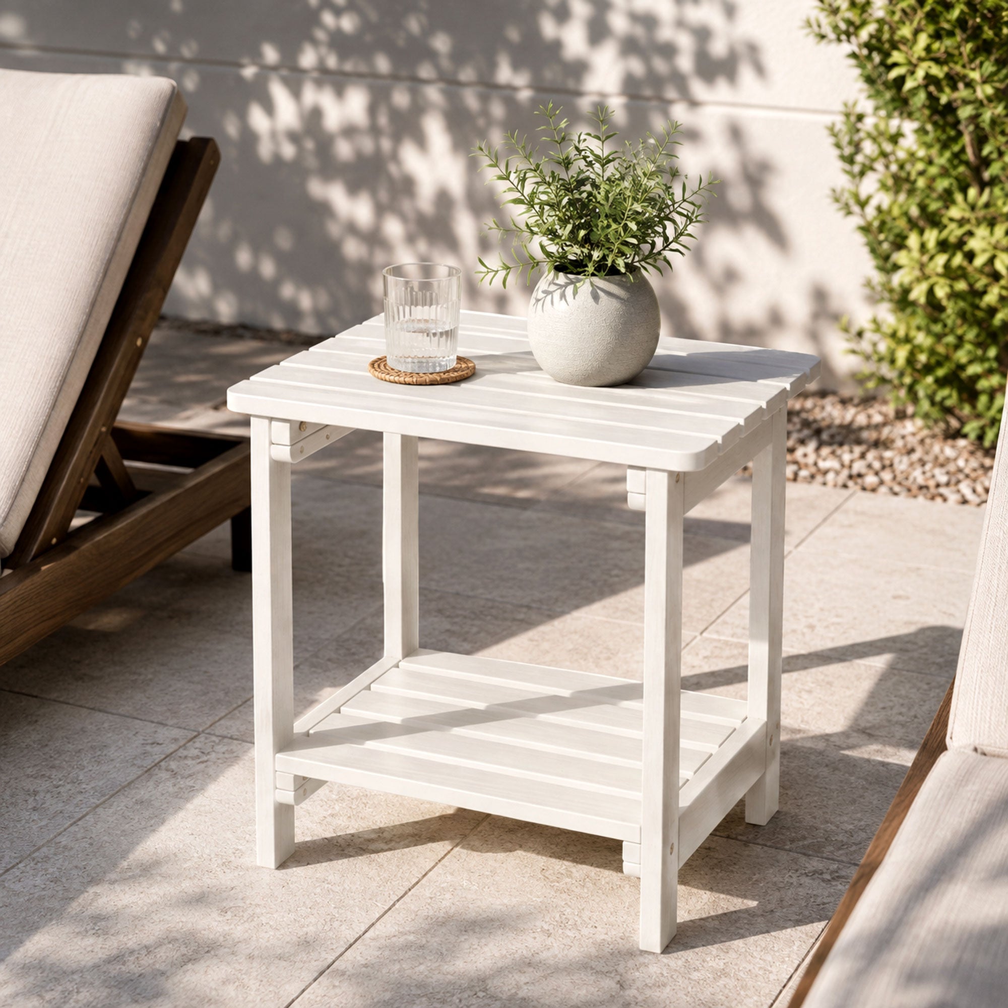 A white outdoor two-tier side table with a slatted top and lower shelf, styled with a small glass and a potted green plant on a patio. A wooden lounge chair and a green shrub are visible in the background. 