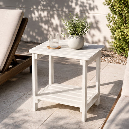 A white outdoor two-tier side table with a slatted top and lower shelf, styled with a small glass and a potted green plant on a patio. A wooden lounge chair and a green shrub are visible in the background. 