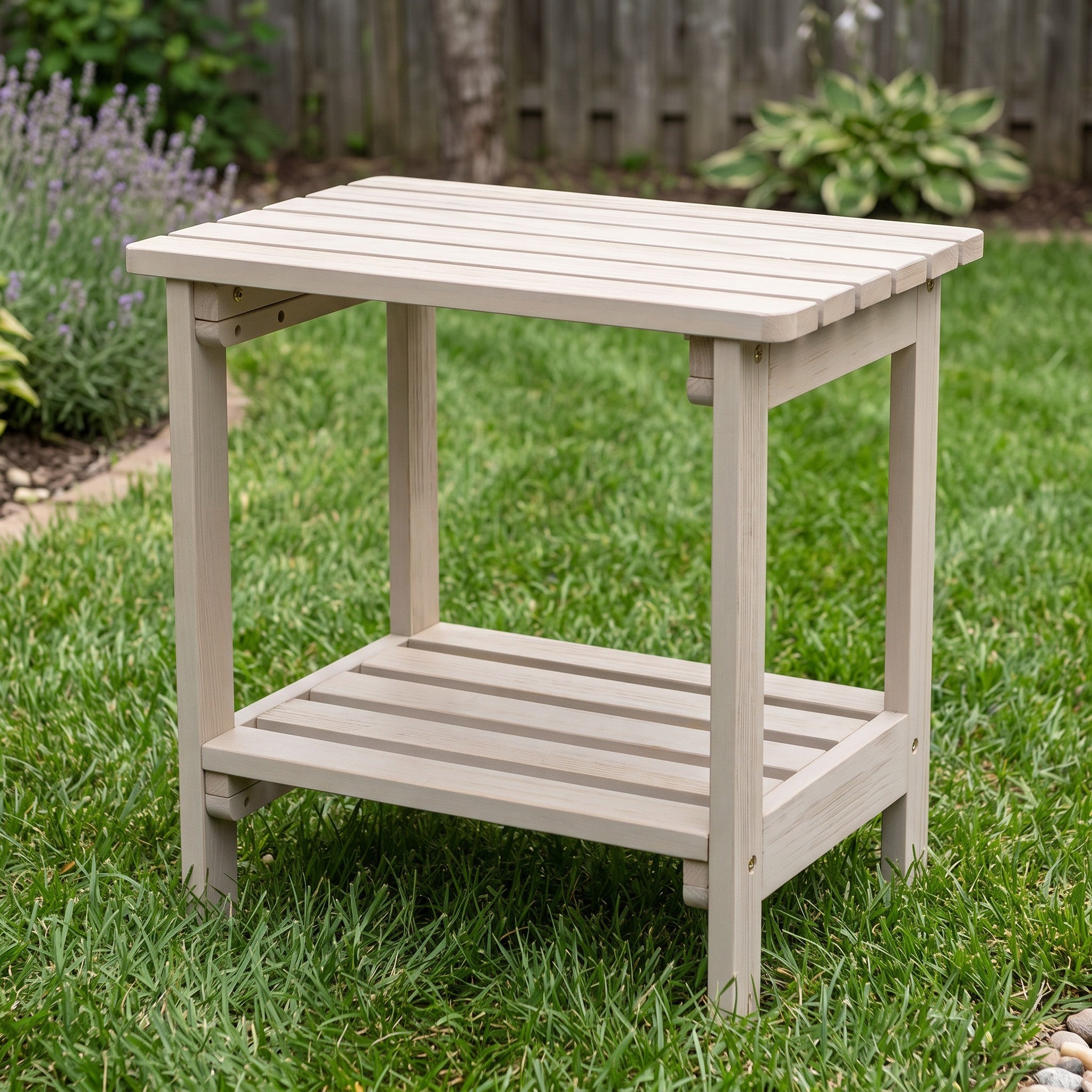 A light beige outdoor wooden end table with a slatted top and lower shelf, shown on a green lawn in a backyard setting. 