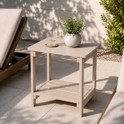 A light gray outdoor side table with a slatted top and lower shelf, styled on a patio with a potted plant and a glass on top. 