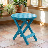 A bright turquoise folding side table with a round slatted top and X-style legs, shown in a sunlit room with tiled floor and potted plants in the background. 