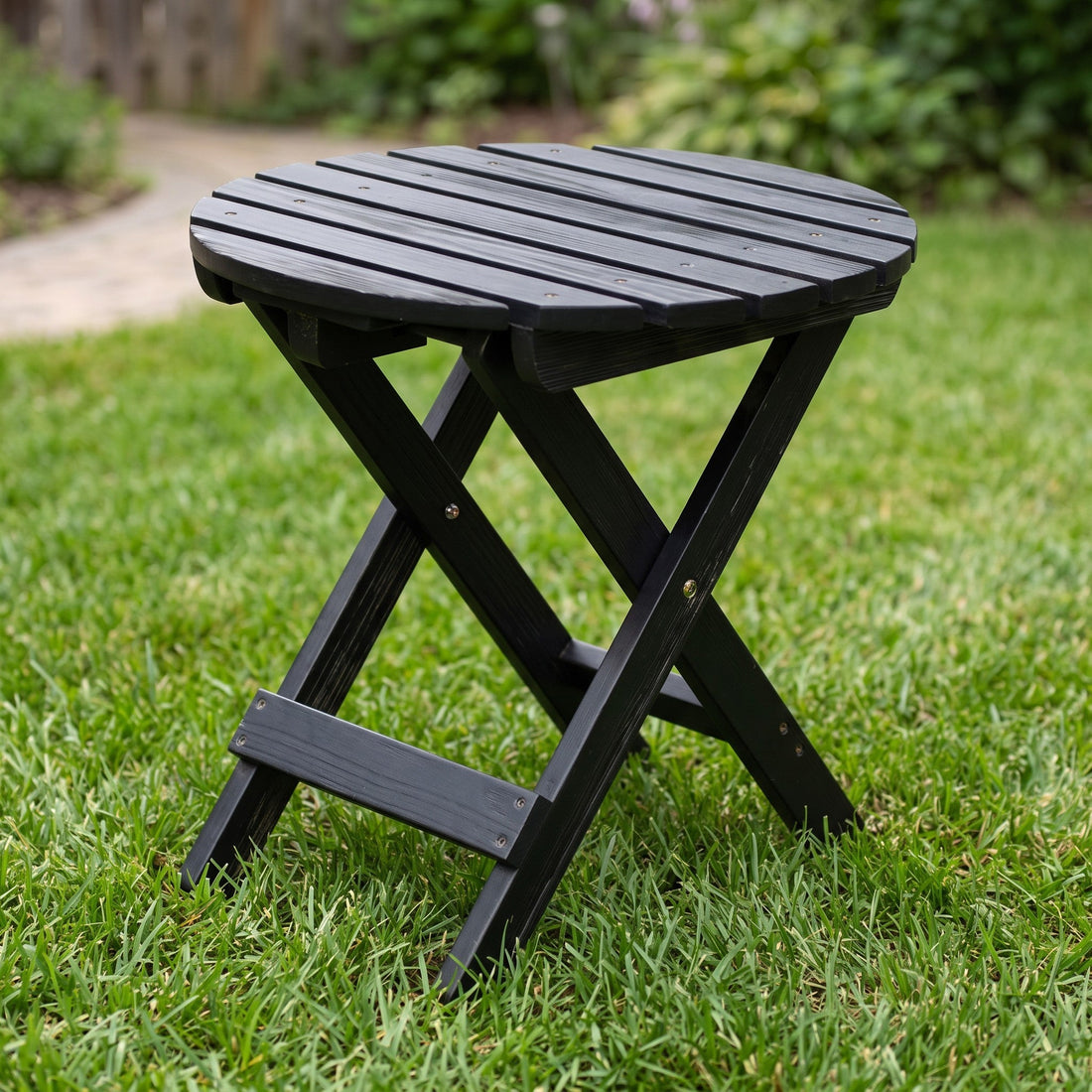 A black folding outdoor table with a slatted round top and X-style legs, shown on a green lawn with a blurred garden background. 