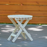A small round folding side table in white, shown outdoors on a gray deck with a wooden fence in the background. 