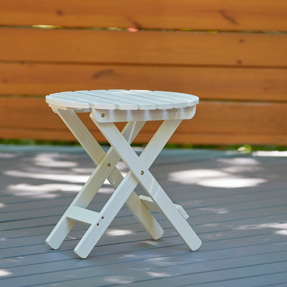 A small round folding side table in white, shown outdoors on a gray deck with a wooden fence in the background. 
