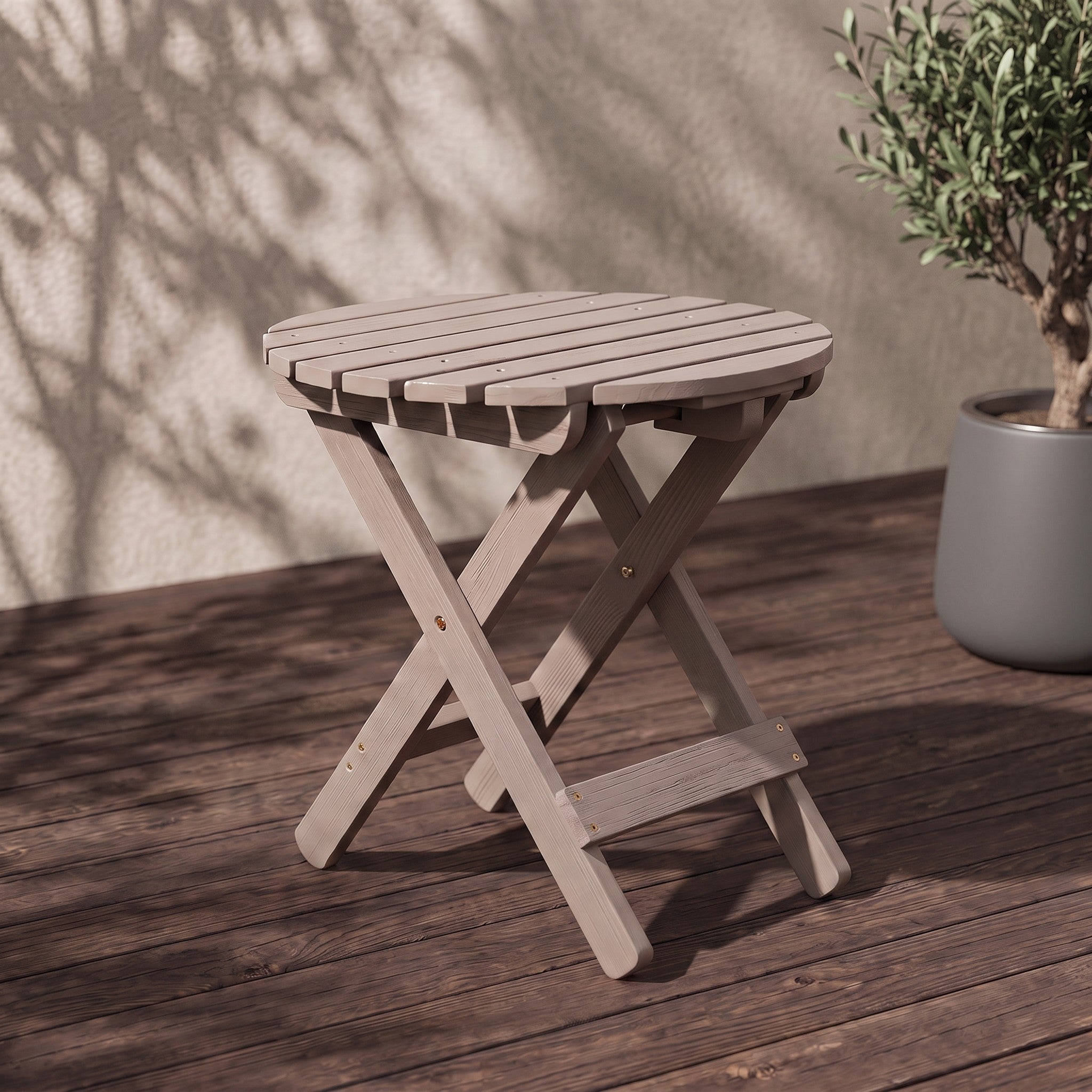 A small round folding stool in a light grayish wood finish, shown on a dark wooden deck with a textured wall and leaf shadows behind it. A potted plant is visible on the right side. 