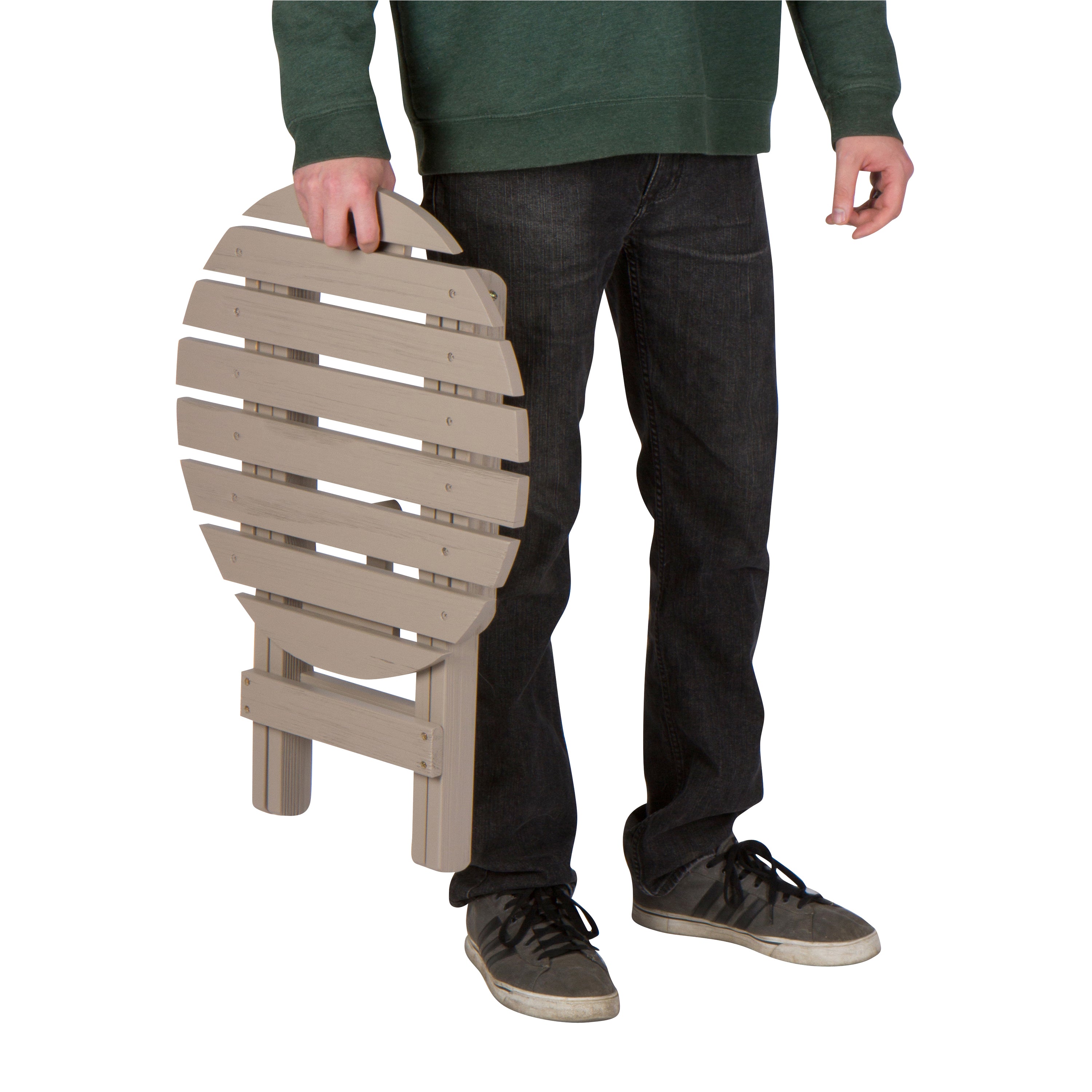 A person wearing a green sweatshirt, dark jeans, and gray sneakers holds a compact, foldable chair made of light beige slatted plastic or composite material against a plain white background. 