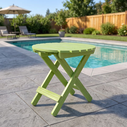 A lime green folding side table with a round slatted top and X-style legs, positioned on a gray stone patio next to a swimming pool in a backyard setting. 