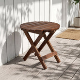 A round folding wooden side table with a slatted top and X-frame legs, shown on a light deck in front of a white wall with dappled sunlight and shadows. 