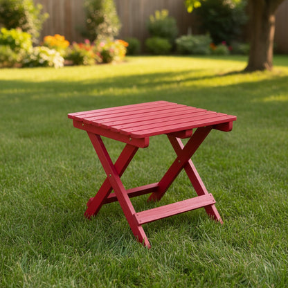A small red folding side table made of slatted wood, set on a green grassy lawn in a backyard with a wooden fence and trees in the background. 