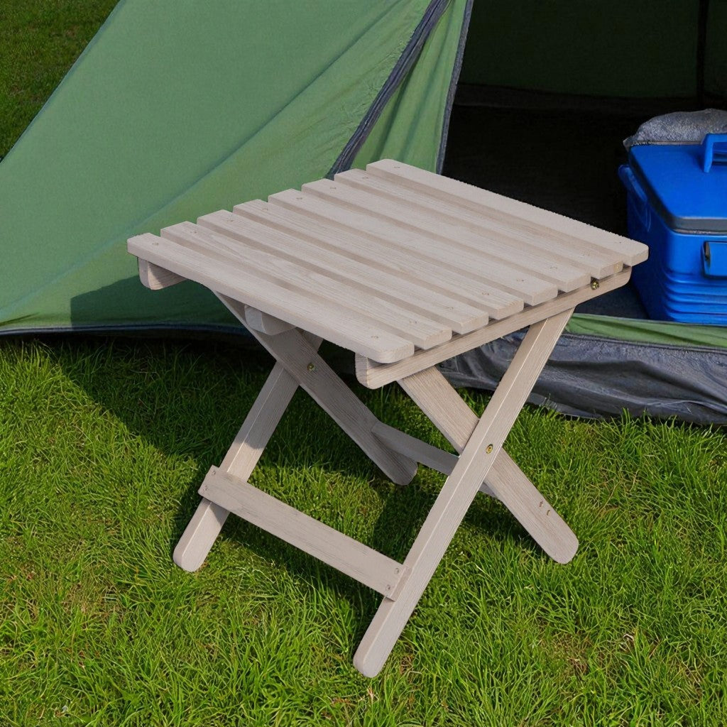 A light-colored folding wooden gray side table with a slatted top, placed on green grass next to a green tent and a blue cooler. 