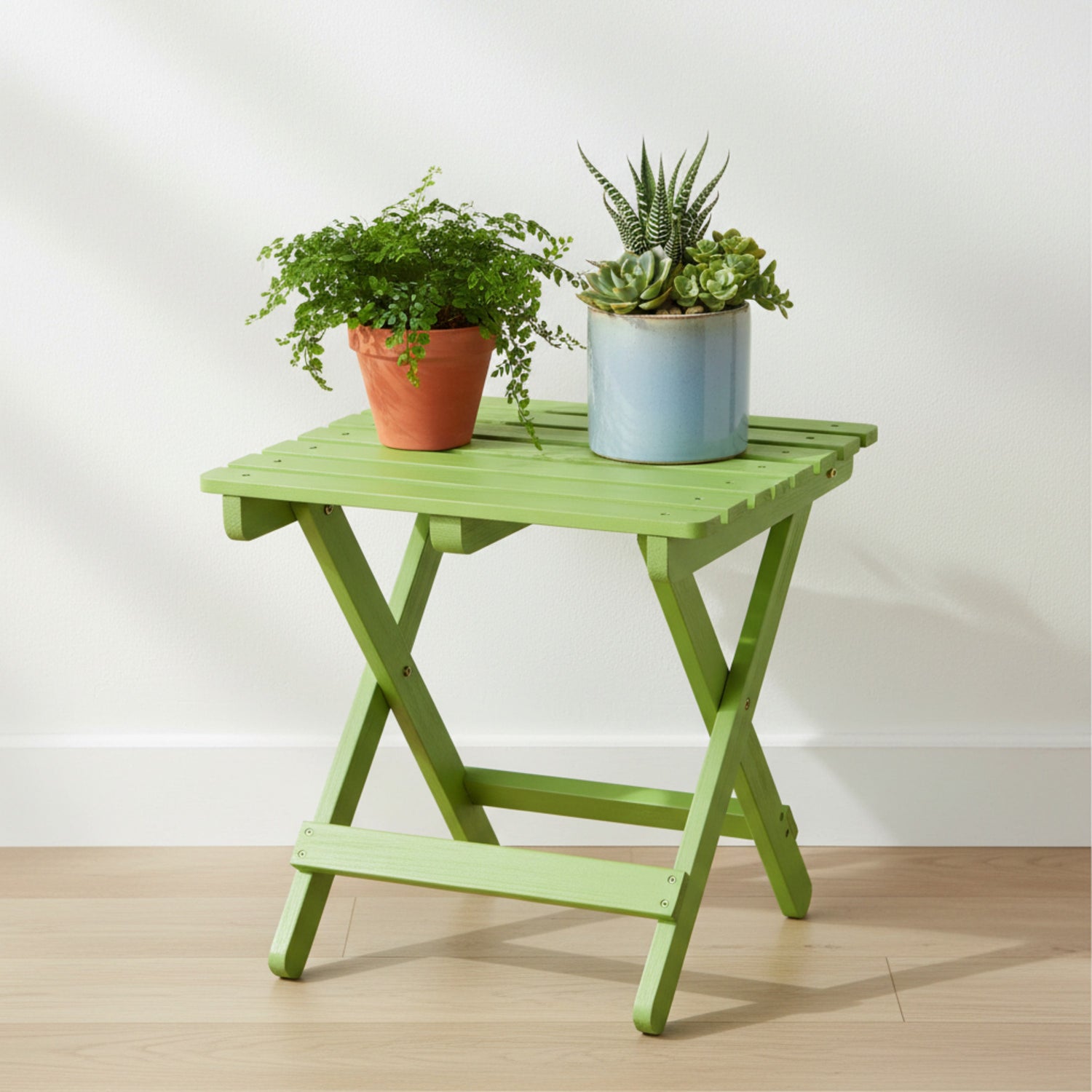 A lime green folding side table with a slatted top and X-frame legs, styled with two potted plants against a white wall and light wood floor. 