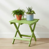 A lime green folding side table with a slatted top and X-frame legs, styled with two potted plants against a white wall and light wood floor. 