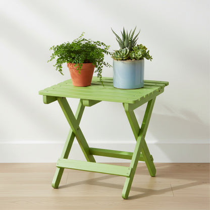 A lime green folding side table with a slatted top and X-frame legs, styled with two potted plants against a white wall and light wood floor. 